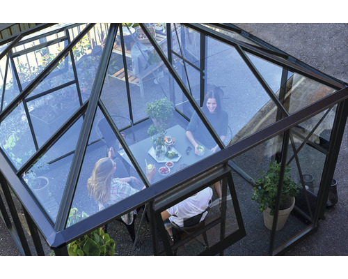 Gewächshaus mit transparenten Wänden und Metallrahmen, im Inneren sitzen Personen an einem Tisch.