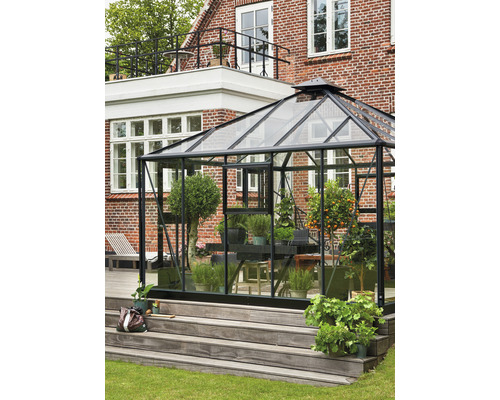 Gewächshaus mit Pflanzen auf einer Holzterrasse vor einem Backsteinhaus.