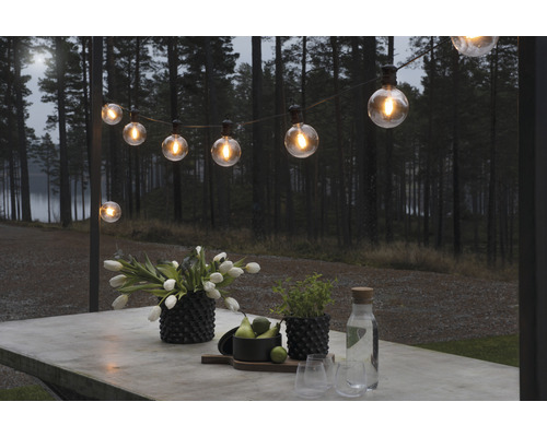 Beleuchtete Lichterkette über einem Tisch im Außenbereich mit Blumen, Pflanzen und Getränken