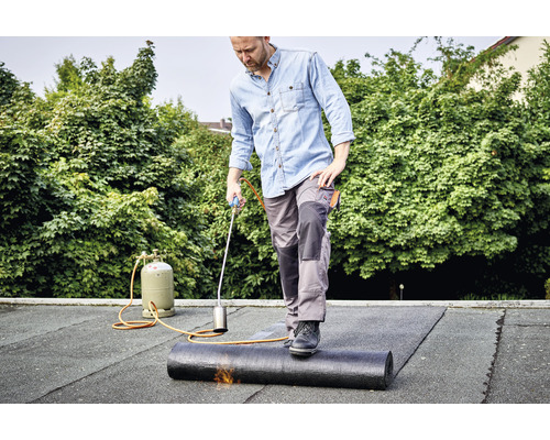 Mann verlegt mit einem Dachbrenner eine Dachbahn auf einem Dach.