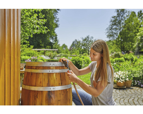 Eine Frau schließt einen Gartenschlauch an ein hölzernes Regenwasserfass im Garten an.