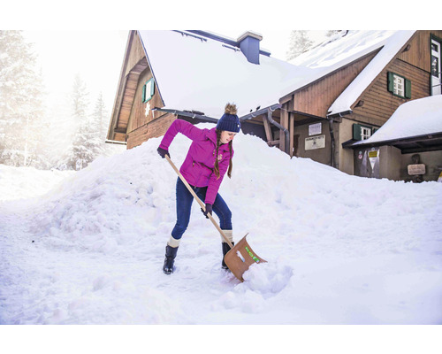 Frau schaufelt Schnee vor einem Holzhaus mit einer Schneeschaufel