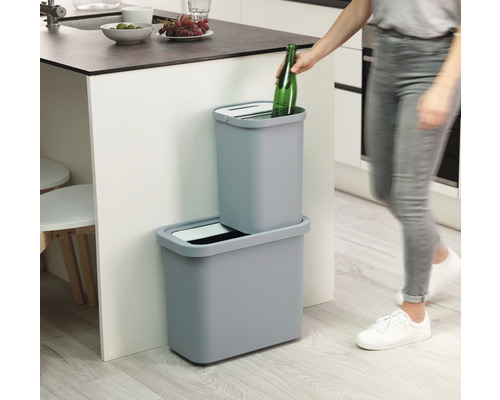 Person wirft eine Glasflasche in einen grauen Recycling-Mülleimer in einer Küche.