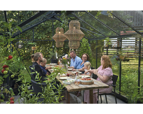 Szene in einem Gewächshaus mit einer Familie, die an einem Holztisch sitzt, umgeben von Pflanzen und zwei hängenden Lampen.