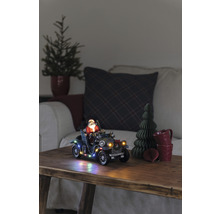 Dekoratives Weihnachtsszenario mit Weihnachtsmann im Oldtimer, beleuchtet, auf einem Holztisch.