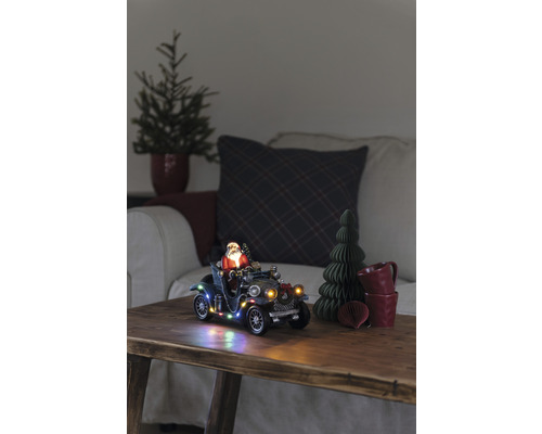 Dekoratives Weihnachtsszenario mit Weihnachtsmann im Oldtimer, beleuchtet, auf einem Holztisch.