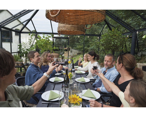 Gruppe von Personen sitzt an einem Tisch in einem Gewächshaus und prostet mit Weingläsern