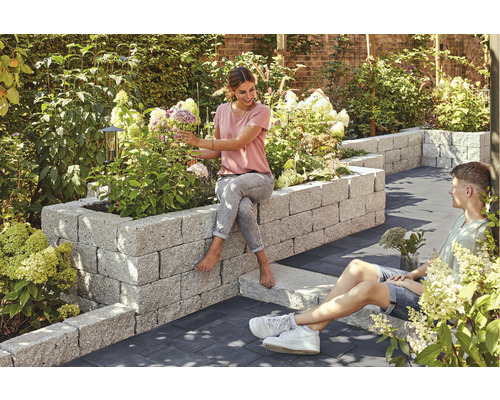 Garten mit Hochbeet aus Stein, bepflanzt mit Blumen, auf dem eine Frau sitzt, während ein Mann daneben auf dem Boden sitzt