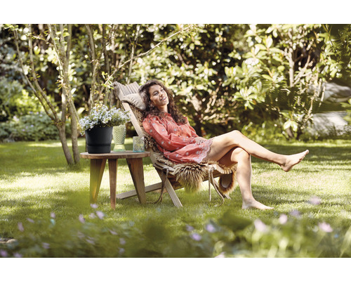 Szene im Garten mit einer Frau, die sich auf einer Holzliege entspannt und neben einem kleinen Tisch mit einem Blumentopf.