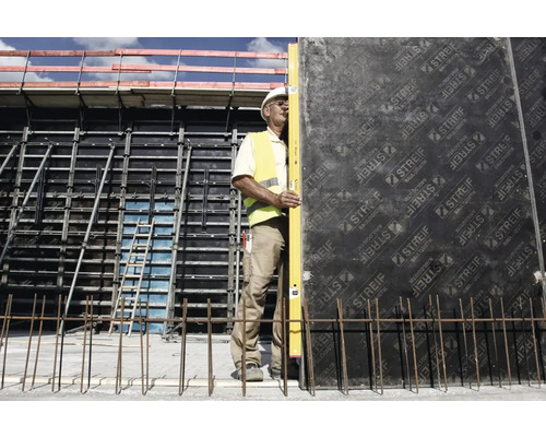 Ein Bauarbeiter mit Helm und Warnweste verwendet eine Wasserwaage an einer Schalung.