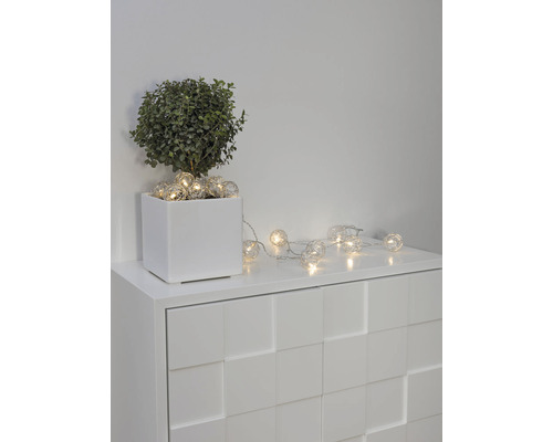 Dekorative Lichterkette mit kugelförmigen Lampen auf einer weißen Kommode neben einer Topfpflanze.