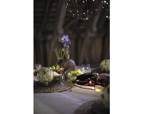 Dekorativer Tisch mit Artischocken, Gläsern, Blumenvase und Lichterketten unter kugelförmigen Lampen.
