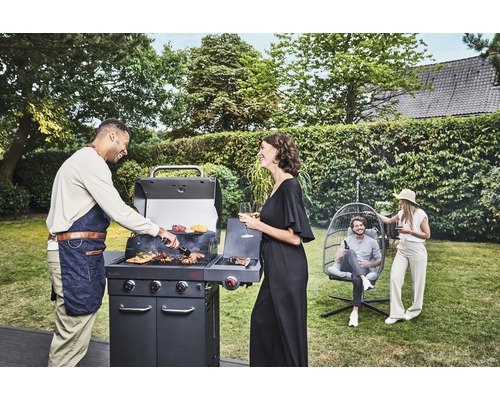 Personen grillen mit einem Gasgrill im Garten, während andere in einem Hängesessel entspannen.