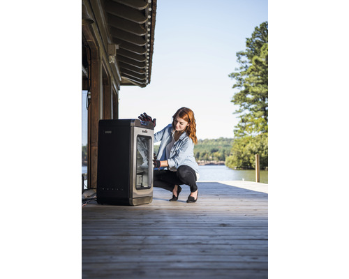 Frau hockt vor einem Räucherofen auf einer Holzterrasse am See