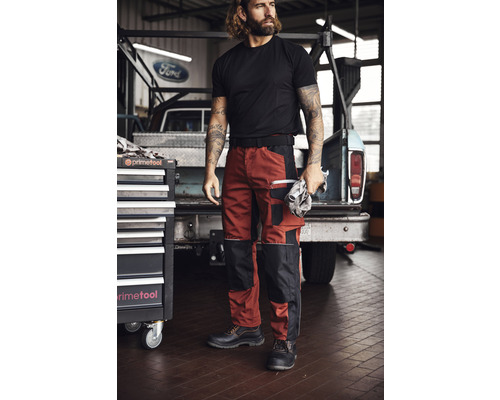 Mann in Arbeitskleidung mit Arbeitshose, T-Shirt und Sicherheitsschuhen vor einem Werkzeugwagen und einem Ford Pickup.