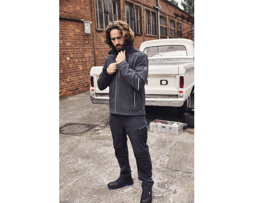 Mann in Arbeitskleidung mit Jacke, Hose und Sicherheitsschuhen vor einem Pickup.