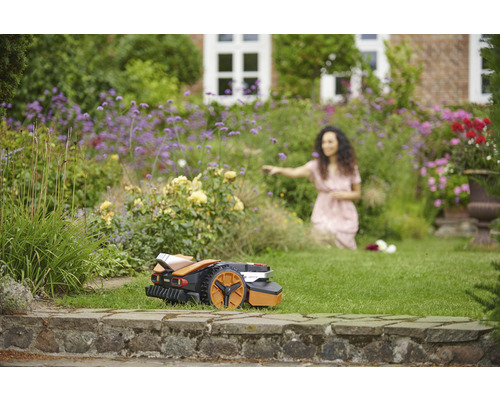 Rasenmähroboter in einem blühenden Garten, während eine Frau sich den Blumen zuwendet
