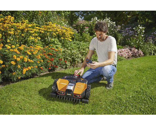 Mann bedient einen Mähroboter mit einem Smartphone im Garten.