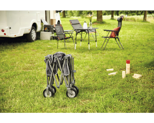 Zusammengeklappter Bollerwagen auf einer Wiese, Campingmöbel und Wohnwagen im Hintergrund.