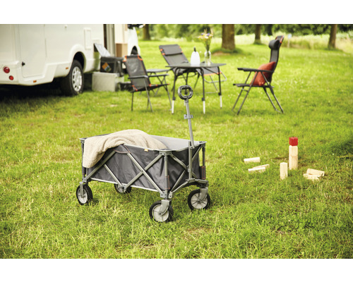 Faltbarer Bollerwagen auf einer Wiese mit Campingmöbeln und einem Wohnmobil im Hintergrund.