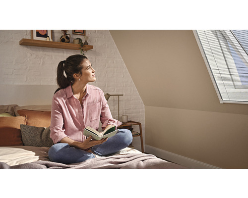 Frau sitzt mit Buch auf einem Bett in einem Schlafzimmer mit Dachfenster und Jalousien.