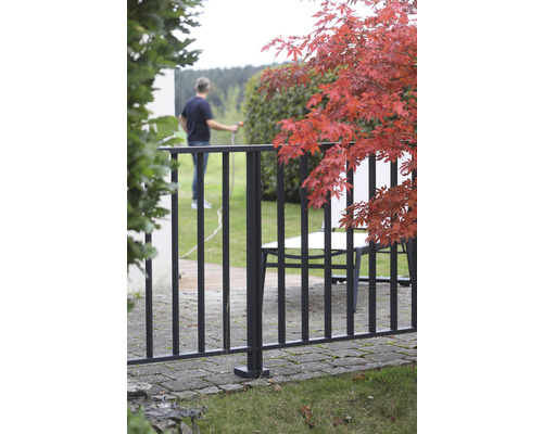 Gartenansicht mit Zaun, Gartenmöbeln und einer Person, die mit einem Gartenschlauch gießt.