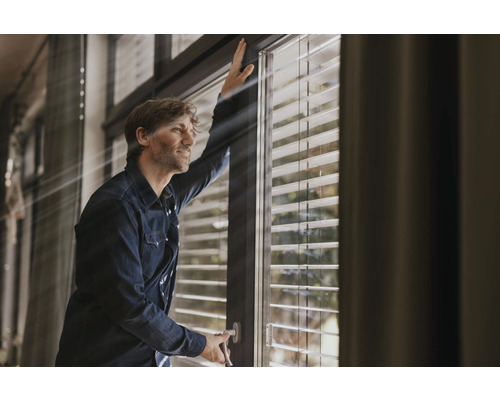 Mann öffnet Fenster mit Jalousien für Tageslicht