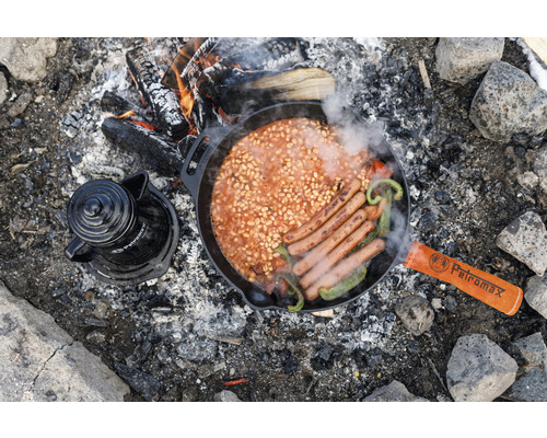 Lagerfeuer-Szene mit Bohnen, Wurst, Paprika in einer gusseisernen Pfanne neben einer Kaffeekanne und dem Petromax Logo