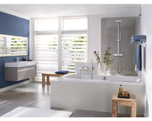 Badezimmer mit Badewanne, Waschbecken, Spiegel und Fenster mit Jalousien für ein helles Ambiente.