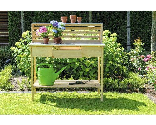 Gartentisch aus Holz mit Ablagefläche, Schublade, Gießkanne, Blumen und Gartengerät im Garten