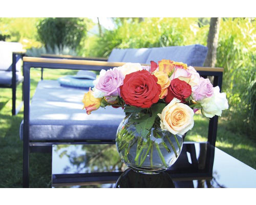 Ein Arrangement aus Rosen in einer Glasvase auf einem Gartentisch mit Gartenmöbeln im Hintergrund.