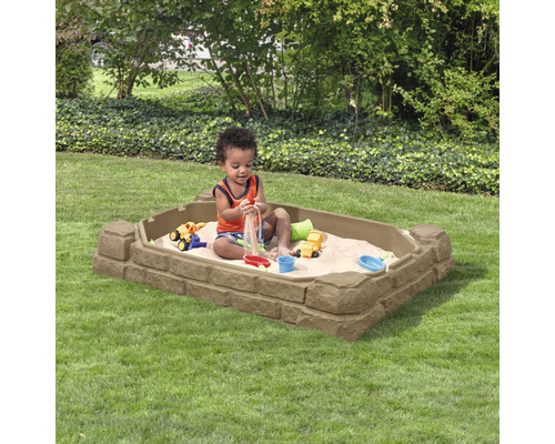 Kleinkind spielt in einem rechteckigen Sandkasten mit Steinmuster im Garten.