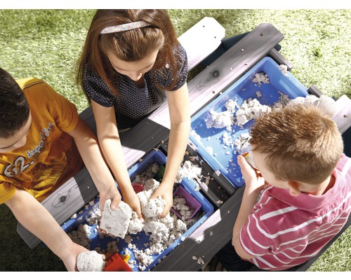 Drei Kinder spielen mit einem Holzsandtisch und Sandspielzeug im Garten.