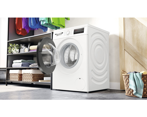 Frontlader-Waschmaschine mit geöffneter Tür in einem Waschraum mit Regalen und Wäschekörben.