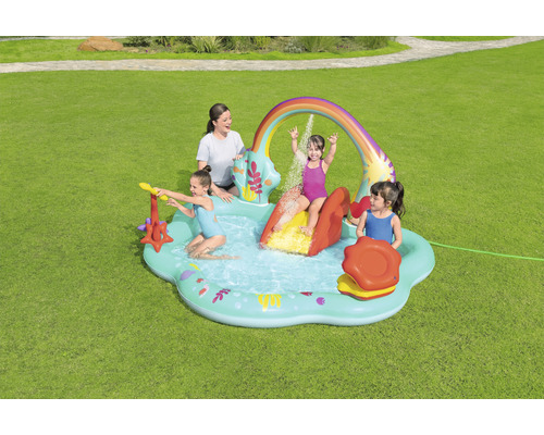 Aufblasbares Planschbecken mit Rutsche, Regenbogen und Wassersprühern im Garten mit spielenden Kindern und Erwachsenen.