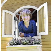 Junge schaut aus dem Fenster eines Holzspielhauses mit Fensterkasten und Bepflanzung