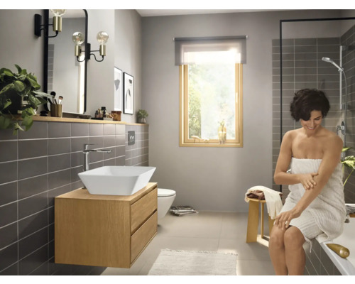 Helles Badezimmer mit Waschtisch, Spiegel, Armatur, Toilette, Dusche und einer Frau mit einem Handtuch.