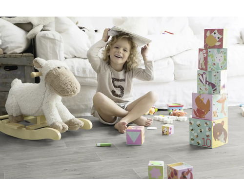 Ein Kind sitzt mit einem Buch auf dem Kopf in einem hellen Zimmer und spielt mit Bauklötzen und einem Schaukeltier.