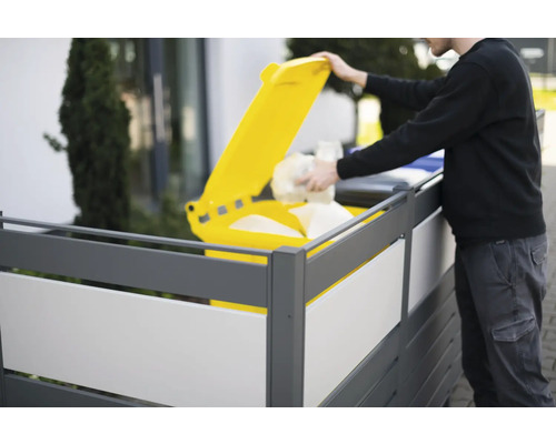 Person wirft Abfall in eine gelbe Mülltonne hinter einem Sichtschutzzaun