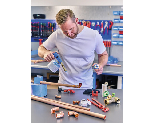 Ein Mann lötet Kupferrohre mit einem Gasbrenner, Lot, Rohren und Fittings auf einer Werkbank.