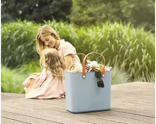 Szene mit einer blauen Tragetasche mit braunen Griffen, gefüllt mit verschiedenen Gegenständen und einer Mutter mit ihrer Tochter im Hintergrund.