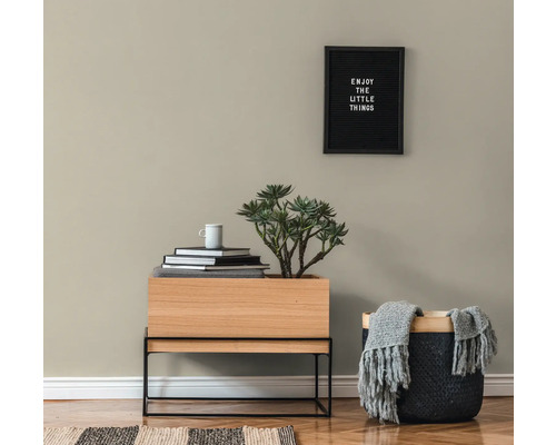 Wohnzimmerszene mit einem Holztisch, einer Pflanze, Büchern, einer Tasse, einem Korb mit einer Decke und einem gerahmten Zitat an der Wand.