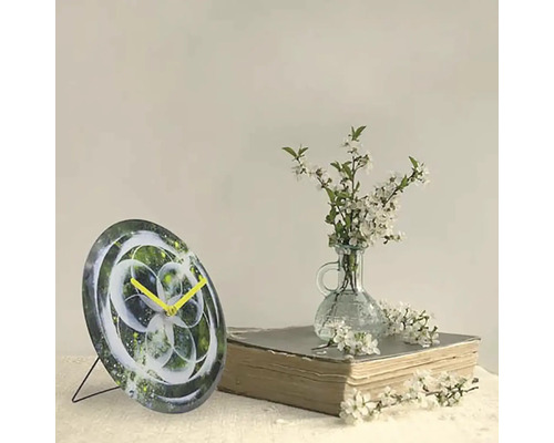 Dekorative Tischuhr mit abstraktem Design und gelben Zeigern neben einem Buch und einer Vase mit weißen Blüten.