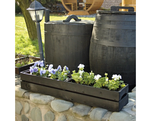 Blumenkasten aus Holz mit Blumen auf einer Steinmauer, im Hintergrund eine Gartenlaterne und Fässer