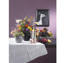 Dekoratives Stillleben mit Blumenarrangement, Obstschale, Kerzen und einem gerahmten Bild von einem weißen Pfau auf einem Tisch mit weißer Tischdecke.