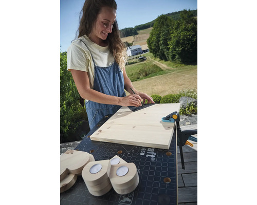 Frau zeichnet Linie auf Holzbrett mit Winkel auf Werkbank mit Schraubzwinge und Holzherzen