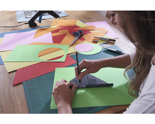 Eine Person zeichnet mit einem Winkel und einem Bleistift auf einem grünen Blatt Papier, umgeben von verschiedenfarbigem Papier, einer Schere und einem Cutter.