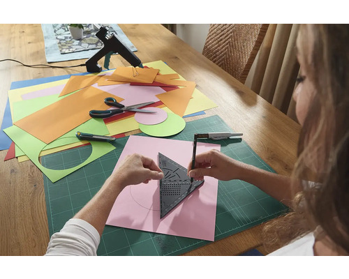 Frau zeichnet einen Kreis mit einem Geodreieck auf ein Stück Papier, umgeben von Bastelmaterialien auf einem Tisch.