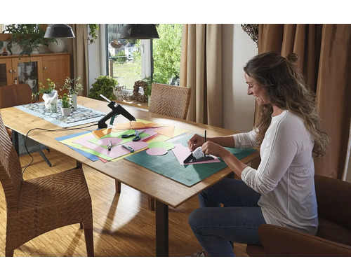 Frau bastelt mit einer Heißklebepistole, Papier, Schere und Schneidematte an einem Tisch