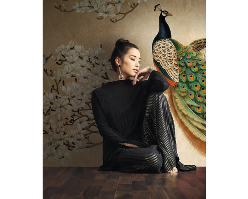 Frau sitzt vor einer Fototapete mit Pfauen- und Blumenmuster.